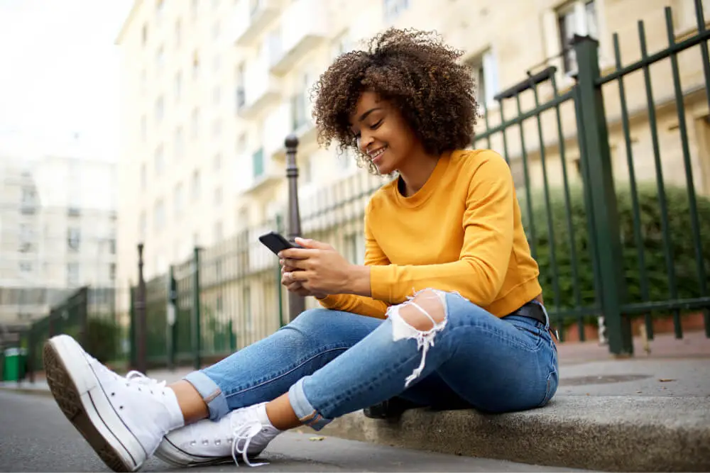 Young woman in casual jeans and sneakers sitting on the sidewalk, looking at her phone and smiling.